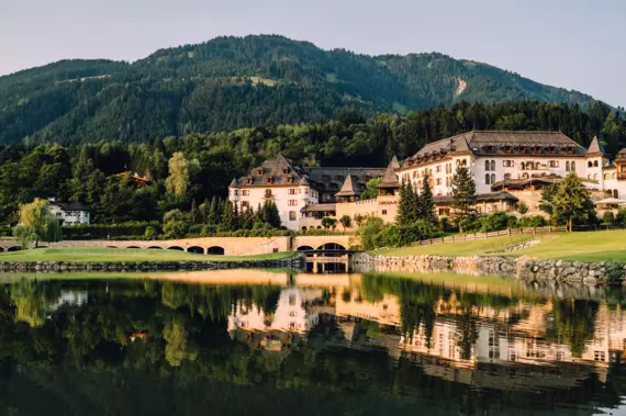 Ein traditionelles Resort spiegelt sich in einem ruhigen See vor der Kulisse eines dicht bewaldeten Berges in der Abenddämmerung.