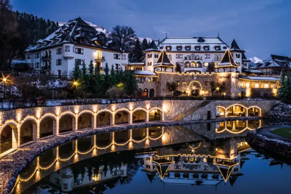 Ein winterlich beleuchtetes Gebäude im alpinen Stil, das A-ROSA Kitzbühel, spiegelt sich in einem ruhigen, künstlichen Teich wider. 