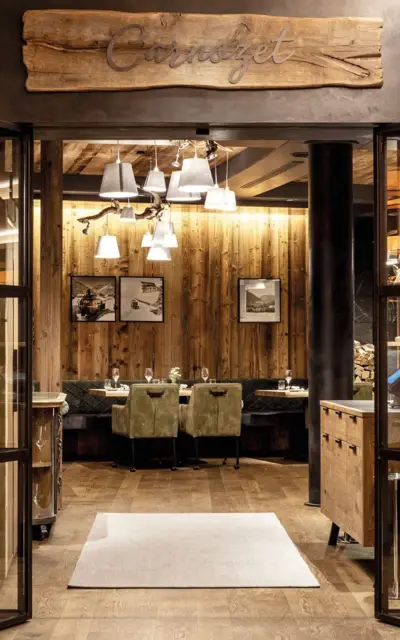 A cozy restaurant area with lots of wooden details in the form of the floor and wooden walls. The chairs and benches are in natural shades of green, pictures hang on the walls and white lampshades hanging from the ceiling provide light.