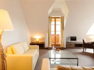 Ein Wohnbereich mit einem hellen Sofa, einem Sessel und einem großen Couchtisch aus Glas. Im Hintergrund ist ein schmales bodentiefes Fenster, ein Fernseher und ein dunkler Sessel zu sehen.