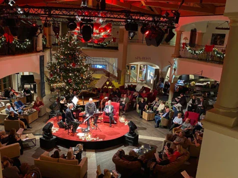 Das Foto zeigt eine belebte Lobby, geschmückt mit festlicher Weihnachtsdekoration. Im Zentrum befindet sich ein großer, prächtig geschmückter Weihnachtsbaum, der von einer Gruppe von Menschen umgeben ist, die sich um eine Live-Band oder Musiker versammelt haben, die auf einer kleinen Bühne spielen. Die roten Akzentlichter beleuchten die sichtbaren Holzbalken der Decke und schaffen eine warme, einladende Atmosphäre. Gäste sitzen auf Sofas und Stühlen, einige halten Getränke in der Hand, während sie der Musik zuhören und die festliche Stimmung genießen.