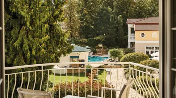 Blick von einem Balkon auf einen Pool, umgeben von Bäumen und einem Gebäude.