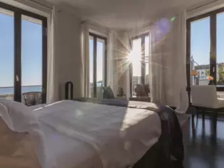 Ein sonnendurchflutetes Schlafzimmer mit vielen großen Fenstern. In der Mitte des Raumes steht ein großes, in weiß bespanntes Bett, mit Blick auf das Meer.  