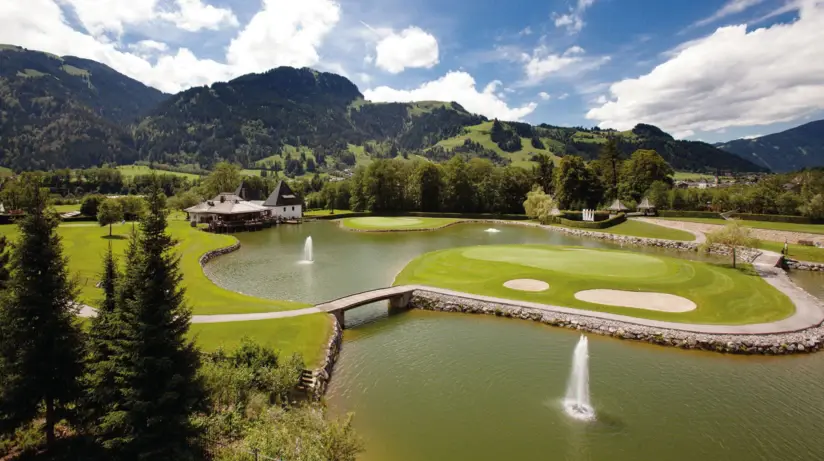 Luftansicht eines Golfplatzes mit gepflegter Grünanlage und großem Teich mit Fontänen, sowie den Bergen im Hintergrund. 