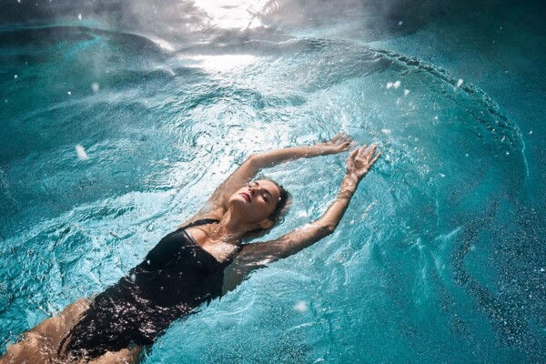 A woman in a black swimsuit floats relaxed on her back in a clear blue pool. She has her arms stretched out above her head and her eyes closed. The water around her forms gentle waves, while sunlight shimmers through the water, creating a calm, relaxing atmosphere.
