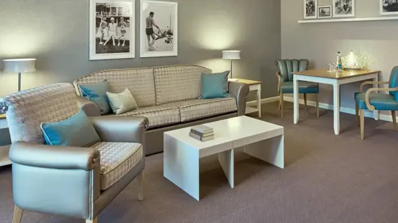 A stylish seating area in a junior suite with a patterned couch in cream and beige tones, accentuated by blue decorative cushions. A modern white coffee table and two matching armchairs complete the seating area. A dining table with green upholstered chairs can be seen in the background. The wall is adorned with attractive gray wallpaper, and framed black and white photographs lend the room a historical flair.