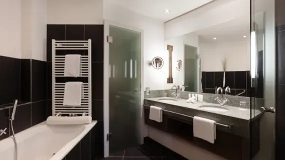 A bathroom with dark tiles, a bathtub and a washstand with two washbasins. A towel warmer with two white towels hangs above the bathtub.