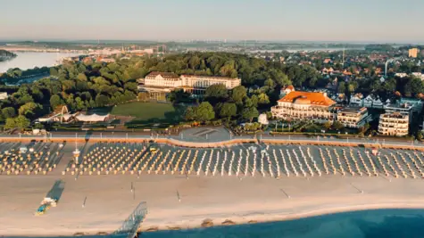 Strand mit vielen Strandkörben und Gebäuden, umgeben von Bäumen und Wasser.