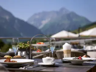 Tisch im Freien mit Geschirr, darunter Teller mit Essen und Getränken mit Bergen im Hintergrund.