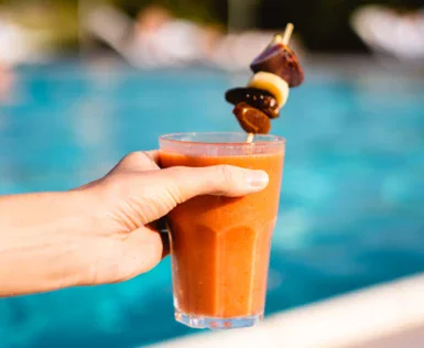 Eine Hand hält einen Smoothie mit Fruchtspieß vor einem unscharfen Hintergrund eines Außenpools mit Sonnenschirmen und entspannenden Gästen.