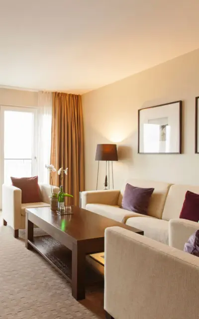 Ein gemütliches Suite-Wohnzimmer in einem Hotel mit natürlichem Licht, ausgestattet mit einer beigen Couchgarnitur mit violetten Kissen, einem dunklen Holztisch und passenden Stühlen. An der Wand hängen zwei gerahmte Landschaftsbilder, und das Zimmer öffnet sich zu einem Balkon mit Stühlen und einem Tisch. Ein Schreibtisch aus dunklem Holz mit einem Flachbildfernseher und eine stehende Lampe vervollständigen die wohnliche Atmosphäre.