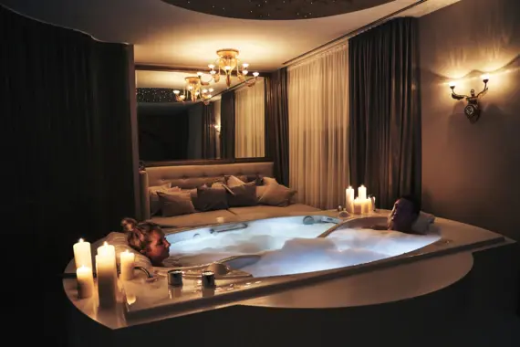 A couple lie in a large, foamy bathtub and enjoy the warmth of the water. Candles stand on the edge of the bathtub, spreading warm light around the room. Behind the bathtub is a comfortable sofa with cushions. 