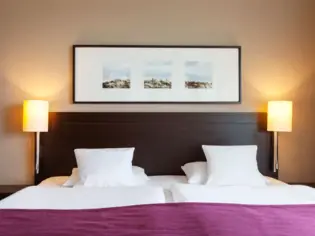 A bed with a dark wood headboard stands in front of a beige wall and a purple bedspread can be seen. An elongated picture frame with three individual square motifs hangs above the headboard.