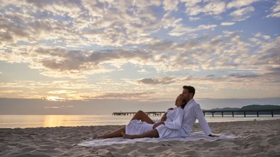 Ein Paar sitzt in Bademänteln bei untergehender Sonne auf einem Handtuch  am ruhigen Strand. 