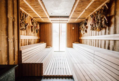 Eine moderne Sauna mit mehrstufigen Holzbänken in einem hellen, natürlichen Holzton. Die Wände sind ebenfalls aus Holz mit einem rustikalen Charakter, und an einer Seite hängen Bündel aus getrockneten Kräutern, die zur entspannenden Atmosphäre beitragen. Über den Bänken ist eine Decke mit sichtbarer Bambusverkleidung. Ein großes Fenster lässt natürliches Licht ein und bietet einen Blick nach draußen.
