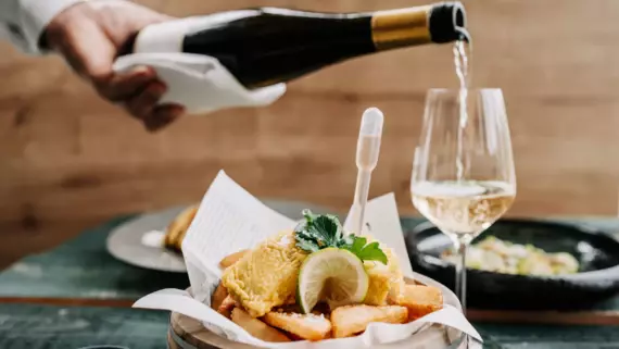 Auf einem Tisch steht ein Teller Fish & Chips und es wird ein Glas Weißwein eingeschenkt.