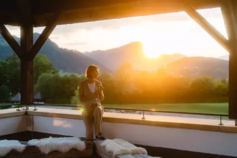  Auf dem Bild ist eine Frau zu sehen, die alleine auf einer Terrasse sitzt und den Sonnenuntergang betrachtet. Sie hält ein Weinglas in der Hand und scheint den Moment der Ruhe zu genießen. Die Szene wird von warmen Sonnenstrahlen beleuchtet, die den Himmel in ein sanftes, goldenes Licht tauchen. Im Hintergrund sind die Umrisse von Bergen zu erkennen, was auf eine idyllische Lage im Alpenraum schließen lässt.