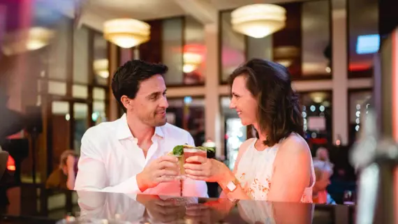 Zwei Personen stoßen in einer belebten Bar mit Cocktails an. Ein Mann in einem weißen Hemd und eine Frau in einem gemusterten Kleid lächeln sich freudig zu, während sie ihre Gläser erheben. Im Hintergrund spiegelt die Bar die leuchtenden Lichter und schafft eine warme und einladende Atmosphäre.