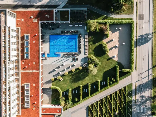 Ein Luftbild zeigt eine Vogelperspektive eines Hotels mit einem großen, rechteckigen, blauen Swimmingpool auf einem begrünten Innenhof. Das rote Dach der Anlage bildet einen auffälligen Kontrast zum umgebenden Grün der gepflegten Rasenflächen und Hecken. 