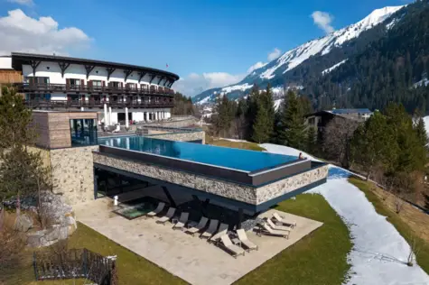 Eine Frau schwimmt in einem ruhigen Innenpool mit vielen Fenstern. Um den Pool stehen hohe, helle Säulen, die von dem warmen Licht gold scheinen. Neben dem Becken sind Nestkörbe und Liegen zur Entspannung bereitgestellt. 