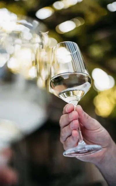 Ein Weiißweinglas wird von einer Hand, vor einem verschwommenen Außenhintergund und einem weiteren Weinglas, in die Luft gehalten. 