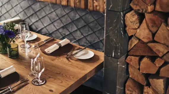 Table with tableware, vase, wine glasses and flowers in front of a wooden wall with firewood stacked on the right edge of the picture.