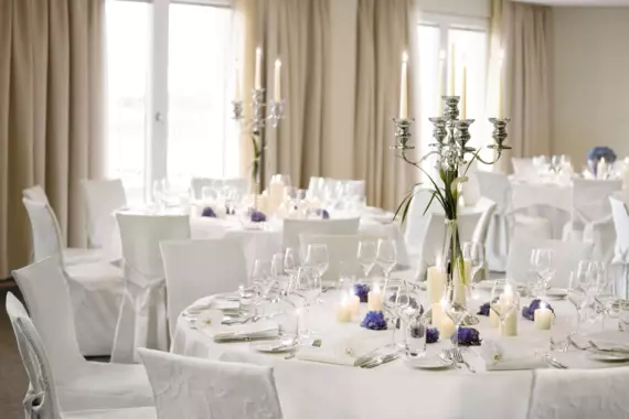 Bestuhlter Hochzeitssaal im A-ROSA Hotel mit Fischgrätparkett, strahlenden Lampen und weißen, runden Tischen mit Hochzeitsdekoration 