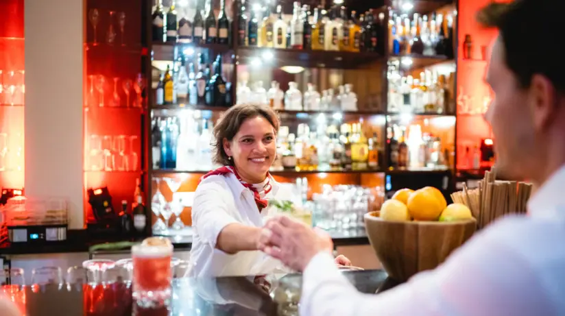 Eine freundliche Barkeeperin reicht einem Kunden in einem belebten Barumfeld einen Cocktail. Sie trägt ein weißes Hemd mit einer roten Krawatte und lächelt, während sie den Drink über den Tresen schiebt. Der Hintergrund der Bar ist mit einer Vielzahl von Spirituosenflaschen beleuchtet.