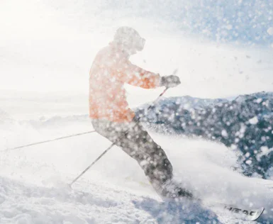 Ein Skifahrer in orangefarbener Kleidung bewegt sich dynamisch durch frischen, aufwirbelnden Schnee bei sonnigem Wetter in einem bergigen Skigebiet