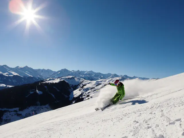  Ein Skifahrer in leuchtend grüner Skibekleidung fährt energisch eine verschneite Piste hinunter, wobei eine Wolke aus aufgewirbeltem Schnee hinter ihm aufsteigt. Die Sonne scheint hell am strahlend blauen Himmel. Im Hintergrund erstreckt sich ein beeindruckendes Panorama von schneebedeckten Bergen und Hügeln.