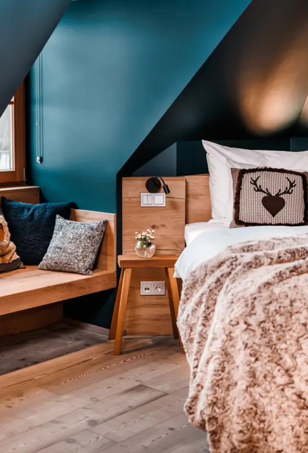 In dem gemütlichen Alpenzimmer steht ein Bett neben einem Nachttisch und einer Fenstersitzbank aus Holz, welche mit kissen verziert ist. Der Raum ist in einem dunklem Türkis gehalten. 