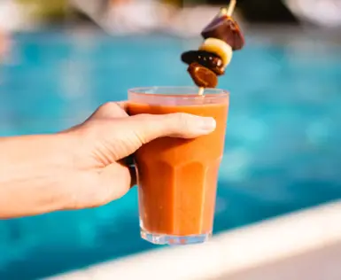 A hand holds a refreshing smoothie with a decorative fruit skewer set against the backdrop of an inviting outdoor pool. The picture radiates summer relaxation and enjoyment, perfect for a day in the wellness area.