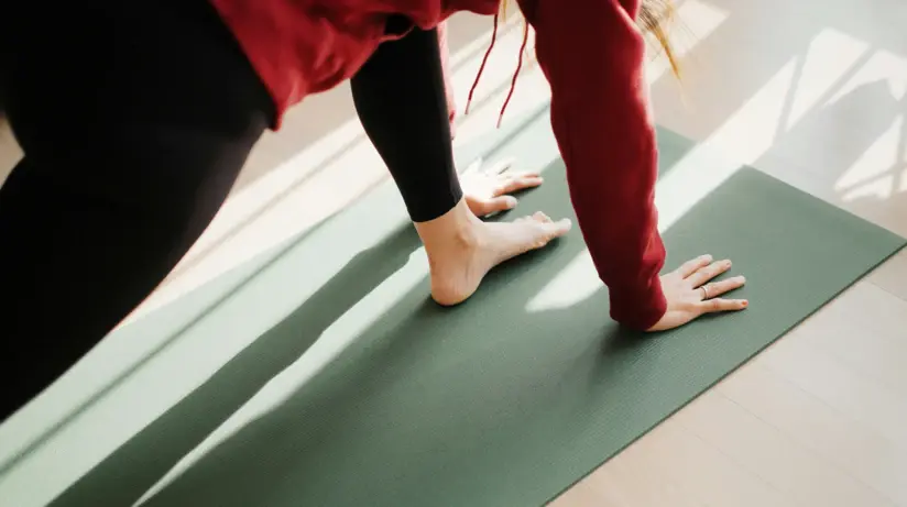 Eine Frau stützt sich mit den Händen auf eine Yogamatte und ihr linken Fuß steht zwischen ihren Händen.