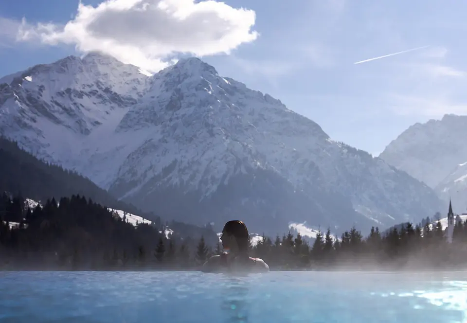 An einem Außenpool von dem A-ROSA Gardasee stehen ordentlich aufgereihte Liegen mit Sonnenschirmen. Im Hintergrund sind Berge und ein Dorf an einem See zu sehen. Die Sonne scheint warm und glitzert auf dem Poolwasser. 