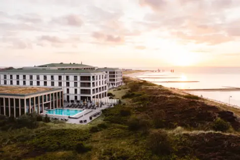 Das Bild zeigt das luxuriöse Resort-Hotel A-ROSA Sylt am Meer während eines ruhigen Sonnenuntergangs. Das sanfte, warme Licht des Abendhimmels taucht die Szene in ein friedliches Ambiente. Die Architektur des Hotels ist modern und elegant, mit klaren Linien und großen Fenstern, die eine offene und einladende Atmosphäre schaffen. Vor dem Hotel befindet sich ein Außenpool, der eine einladende Erfrischung verspricht. Die Dünenlandschaft im Vordergrund mit ihren sanften Hügeln und dem robusten Küstengras bildet einen natürlichen Kontrast zur strukturierten Menschenhand des Hotels. Im Hintergrund ist die weite, ruhige Nordsee zu sehen, die in das warme Glühen des Sonnenaufgangs übergeht und ein Gefühl von Ruhe und Ausgeglichenheit vermittelt.