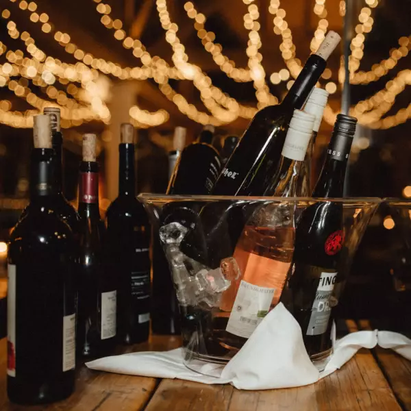  Auf dem Bild ist ein großer, transparenter Eiskübel zu sehen, der mit verschiedenen Flaschen Wein und Sekt gefüllt ist. Einige Flaschen stehen aufrecht, während andere schräg im Eis liegen. Im Hintergrund sind verschwommene Lichterketten, die eine festliche und gemütliche Atmosphäre erzeugen. Die Szene könnte sich auf einer Abendveranstaltung, einer Feier oder in einem stilvollen Restaurant befinden.