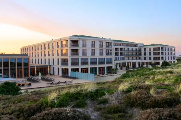 Das Bild zeigt eine Luftaufnahme eines großen, modernen Hotels auf Sylt am Strand. Das Hotel ist von Dünenlandschaften umgeben.  Im Vordergrund führt eine Treppe durch die Dünen, und im Hintergrund ist das ruhige Meer zu sehen. Das klare Wetter und der blaue Himmel verleihen dem Foto eine ruhige, erholsame Atmosphäre.