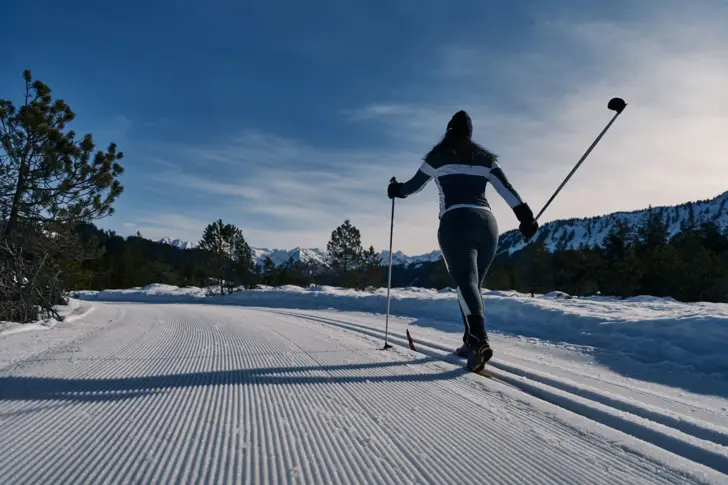 Skilangläuferin auf verschneiter Straße vor einer Bergkulisse.