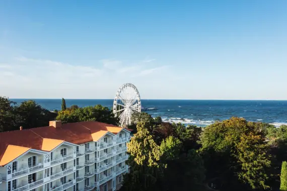 Großes Hotelgebäude im Stil der Bäderarchitektur mit rotem Dach zwischen grünen Bäumen und einem weißen Riesenrad und dem Meer im Hintergrund.