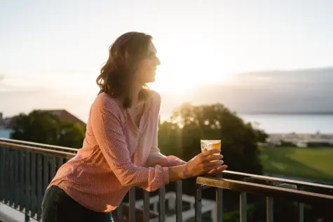 Eine Frau steht bei Sonnenaufgang über ein Balkongeländer gestützt. Lächelnd hält sie ein Glas in ihrer Hand und schaut in die Ferne. Im Hintergrund erstreckt sich eine grüne Wiese und über dem angrenzenden Meer geht die Sonne auf.