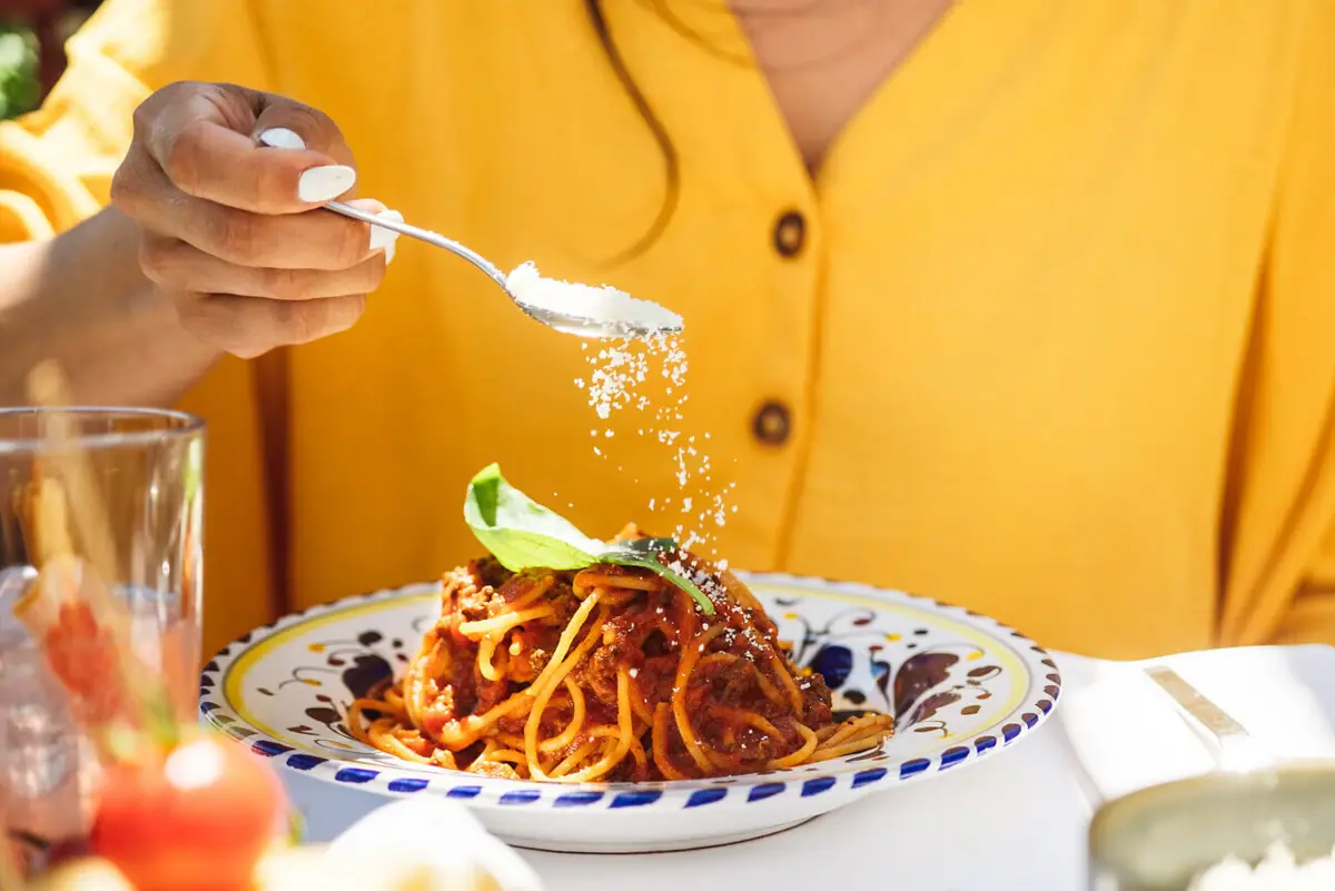 Eine Person streut Parmesan auf einen Teller Spaghetti.
