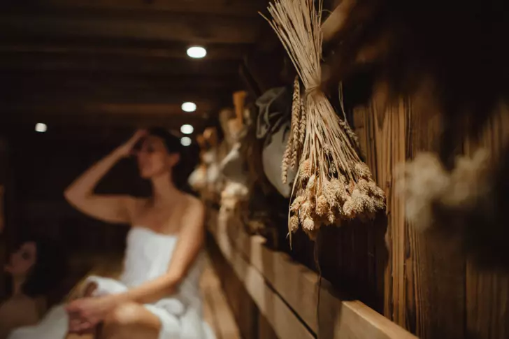 Eine Frau sitzt entspannt in einer Sauna. Sie hat ein Handtuch um den Körper geschlungen und fasst sich in das Haar. Im Fokus steht ein Bündel von Getreideähren, das an der holzverkleideten Wand hängt.