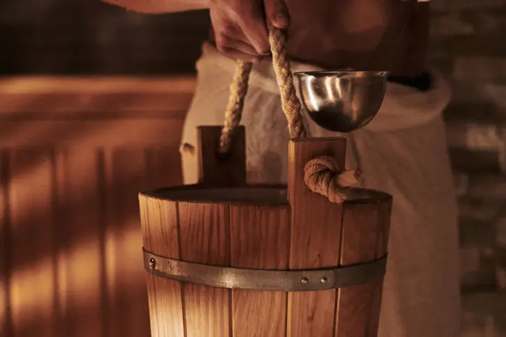 Close-up of a hand holding a wooden sauna bucket, equipped with a metal ladle. The bucket is held by a sturdy rope, which conveys an authentic sauna atmosphere. The scene takes place in warm, subdued light, which emphasizes the woody interior of a sauna and alludes to the traditional infusion ritual in a wellness area.