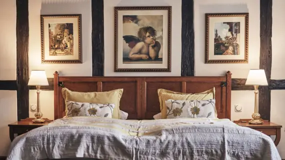 Gemütliches Doppelzimmer im Gotischen Haus Wernigerode mit rustikalem Holzbett, dekorativen Kissen, warmem Licht und kunstvollen Gemälden über dem Kopfteil.