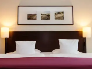 Ein Bett mit einer lila Tagesdecke und einem dunklen Kopfteil steht vor einer hellen Wand. An dieser hängt ein länglicher Bilderrahmen mit drei quadratischen Motiven.