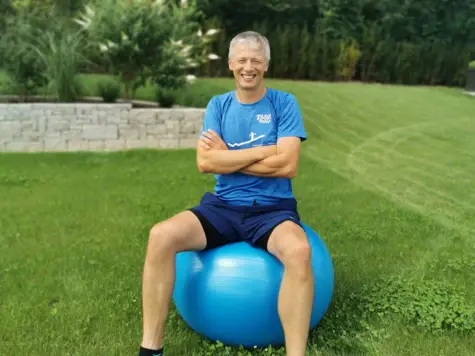 Ein lächelnder Mann sitzt in Sportkleidung mit verschränkten Armen auf einem blauen Medizinball. 