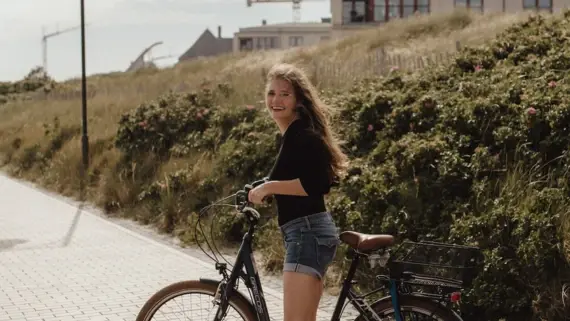 Eine Frau steht lächelnd mit einem Fahrrad auf einem gepflasterten Weg. Im Hintergrund erstreckt sich eine Dünnen Landschaft, welche in sommerliches Licht getaucht ist. 