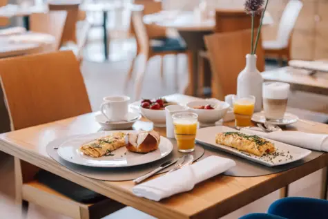 Ein gedeckter Frühstückstisch in einem hellen Restaurant. Auf zwei Tellern sind mit Kräutern bestreute Rühreier angerichtet, es stehen Gläser mit Orangensaft und jeweils ein Kaffee neben den Tellern. 