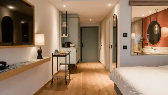 Modern and inviting hotel room with harmonious lighting. On the right in the picture, a large bed with white bed linen in front of a large window with a view of the bathroom, where round mirror lighting and warm colors dominate. On the left is a wooden sideboard with a stylish floor lamp on it, next to a decorative serving trolley with decorative elements on it. The entrance area of the room can be seen in the background.