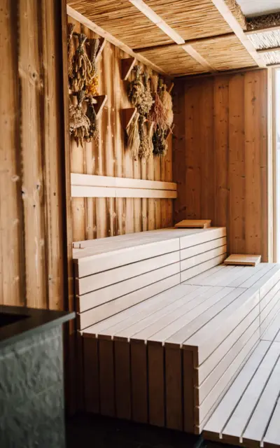 Inneres einer Sauna mit traditioneller Holzverkleidung an Wänden und einer Reetverkleidung an der Decke. Die mehrstufigen, hellen Saunabänke aus Holz sind für mehrere Personen ausgelegt. An der Wand hängen Bündel aus getrockneten Kräutern, die eine natürliche Dekoration darstellen und für ein authentisches Saunaerlebnis sorgen. Natürliches Licht scheint durch ein Fenster herein und betont die warme Atmosphäre des Raumes.